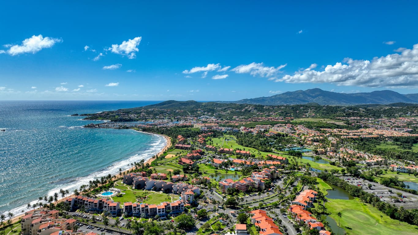 Aerial view of Palmas del Mar, Puerto Rico: a coastal resort lush greenery, golf courses, and the coastline meeting the blue ocean, surrounded by mountains under a bright blue sky.